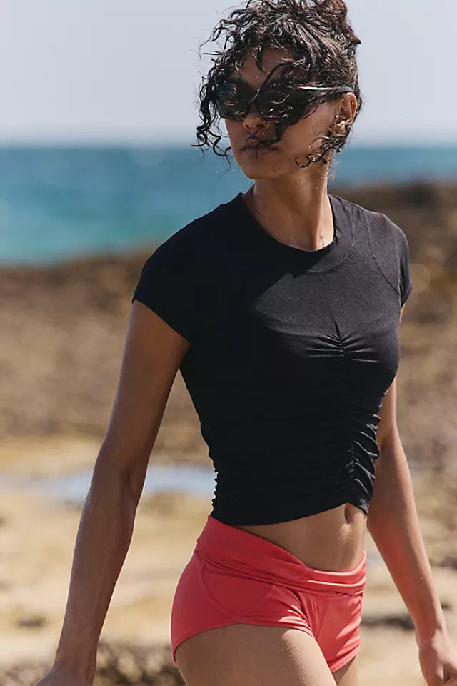 A woman walking on a beach wearing a black short-sleeve ruched crop top and coral pink swim shorts, with sunglasses and a blurred ocean background.