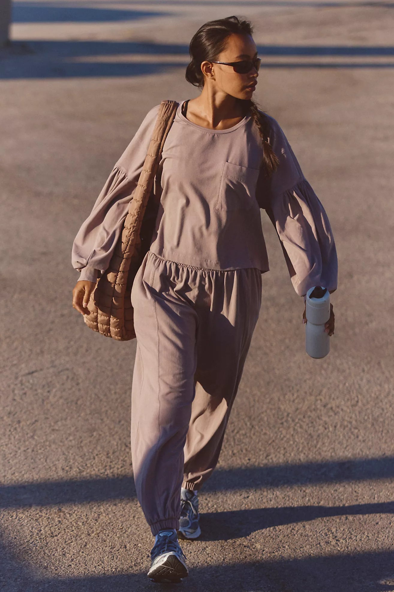 A woman walking outdoors in a matching dusty mauve lounge set, featuring a long-sleeve top and jogger pants. She is carrying a brown woven shoulder bag and holding a white bottle.