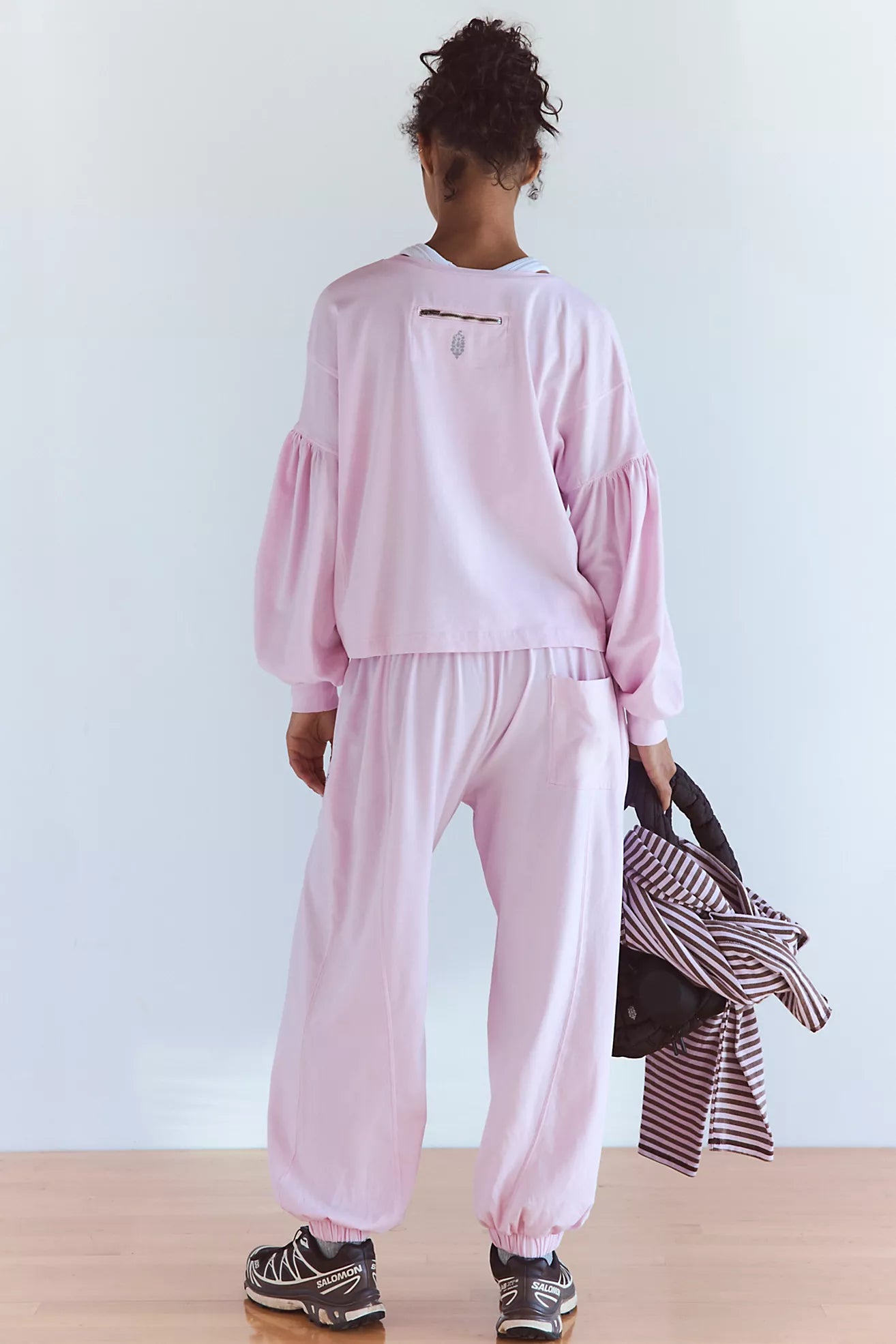 A model shown from behind wearing a light pink two-piece lounge set consisting of a long-sleeve sweatshirt with balloon sleeves and matching jogger pants. The model is holding a striped shirt and a black bag against a clean white wall and light wood floor.