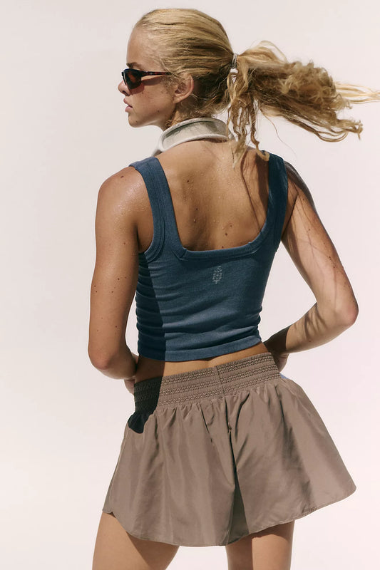 A woman shown from the back wearing a dark blue ribbed scoop-neck athletic tank top and a taupe-brown pleated athletic skirt, set against a plain light background.