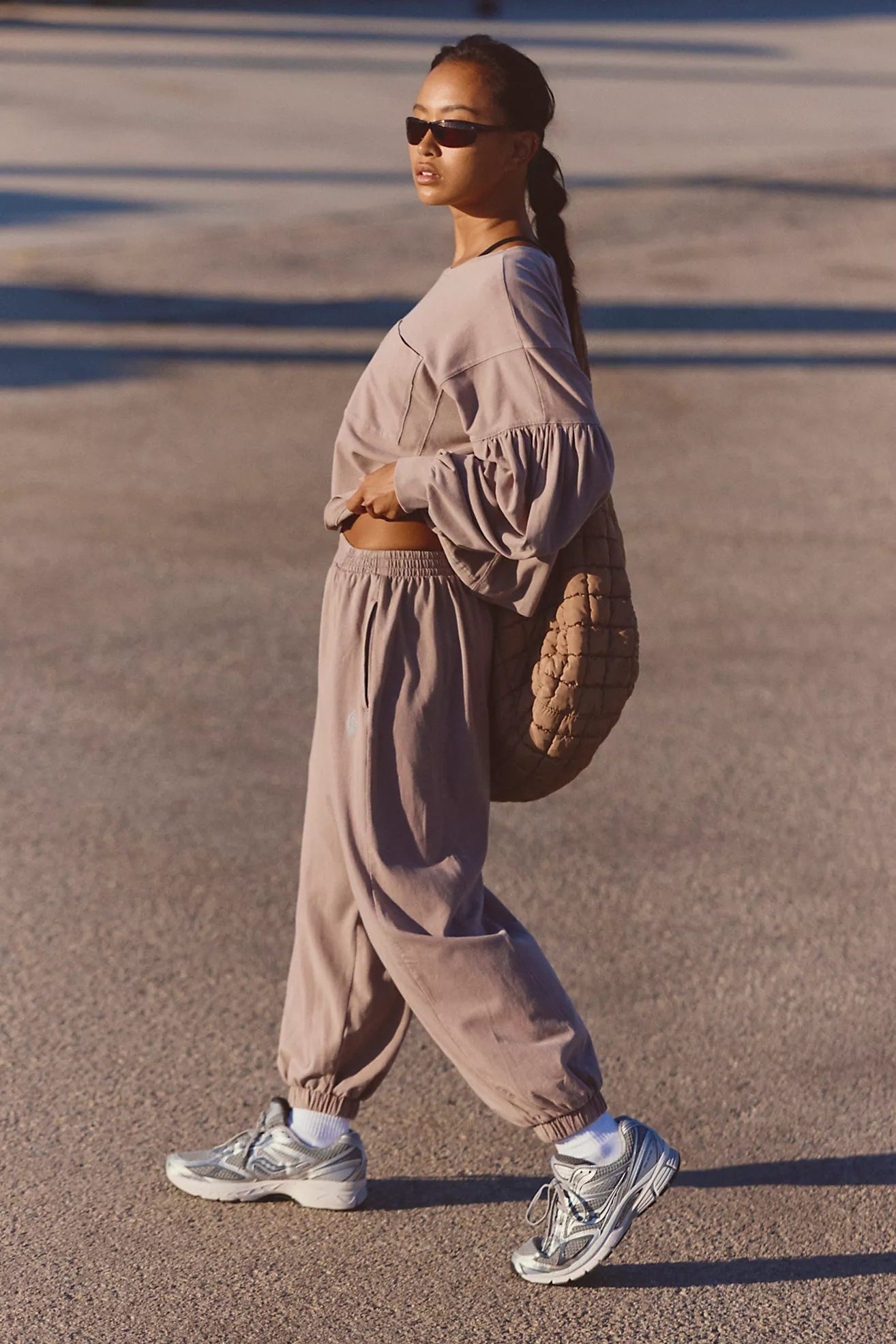 A woman walking outdoors in a matching dusty mauve lounge set, featuring a cropped long-sleeve top and jogger pants, paired with silver sneakers and a brown quilted shoulder bag.