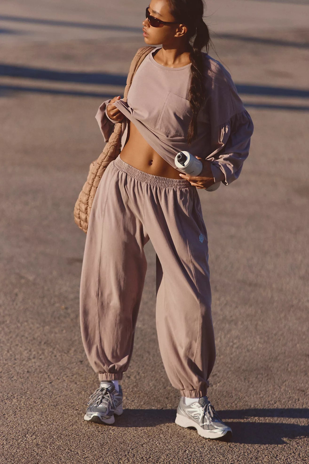 A woman wearing a matching dusty mauve activewear set consisting of a cropped long-sleeve top and loose jogger sweatpants, styled with a beige quilted shoulder bag and silver sneakers on an outdoor paved surface.
