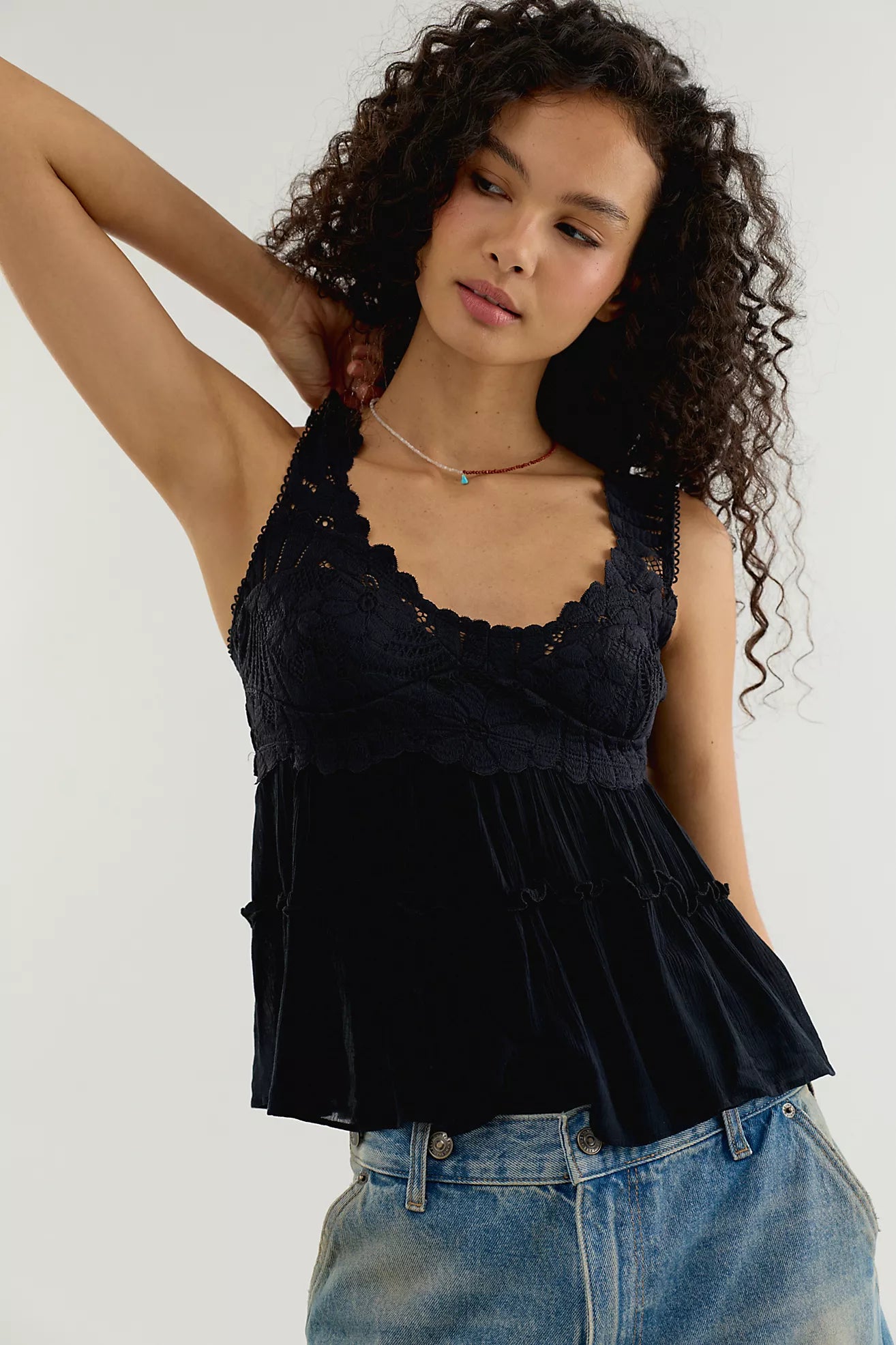 A woman wearing a black lace and velvet camisole top paired with blue jeans, posing against a light gray studio background.