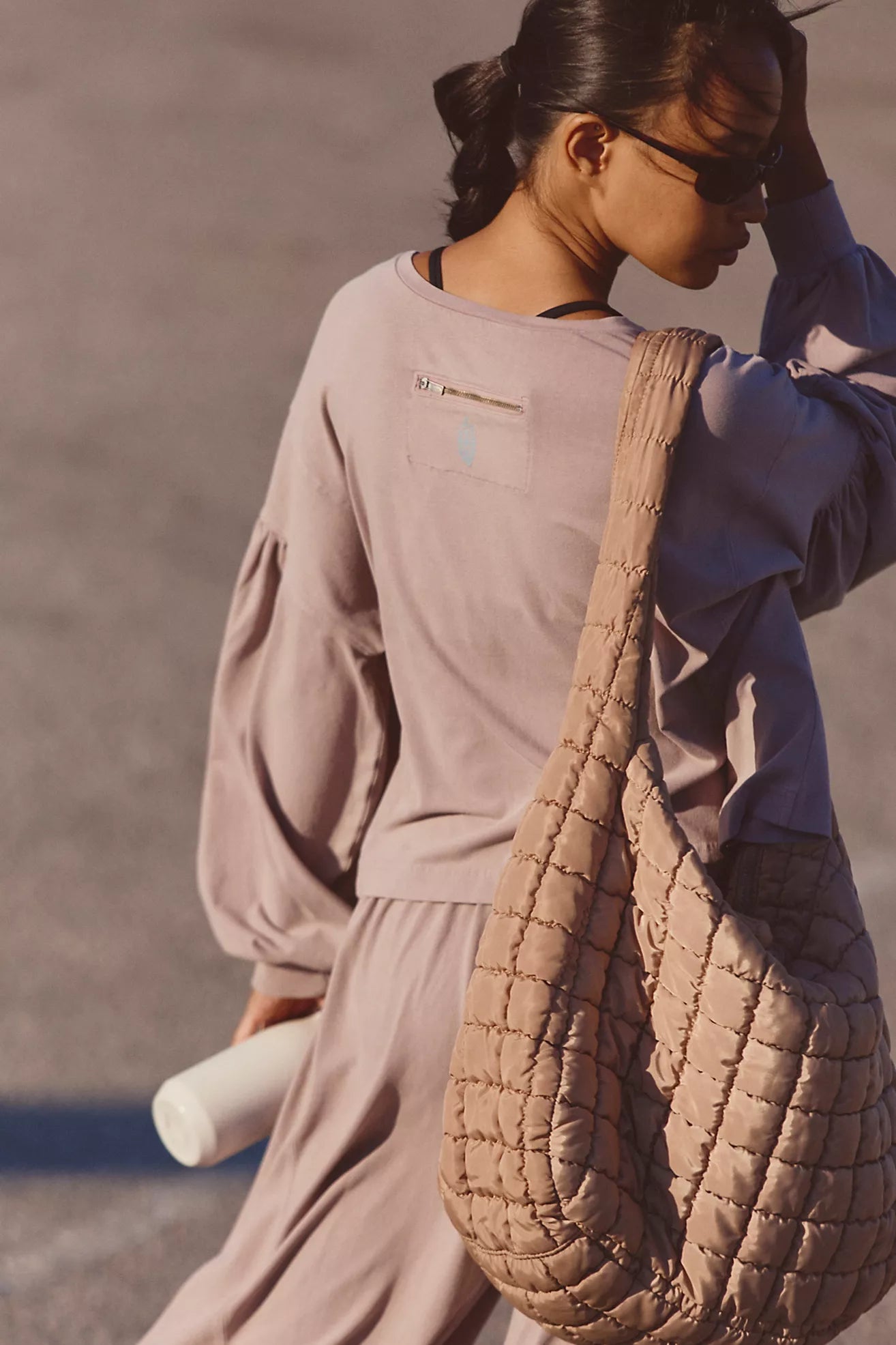 A woman wearing a dusty mauve long-sleeve top and matching skirt, carrying a large quilted taupe shoulder bag, photographed outdoors in warm sunlight.