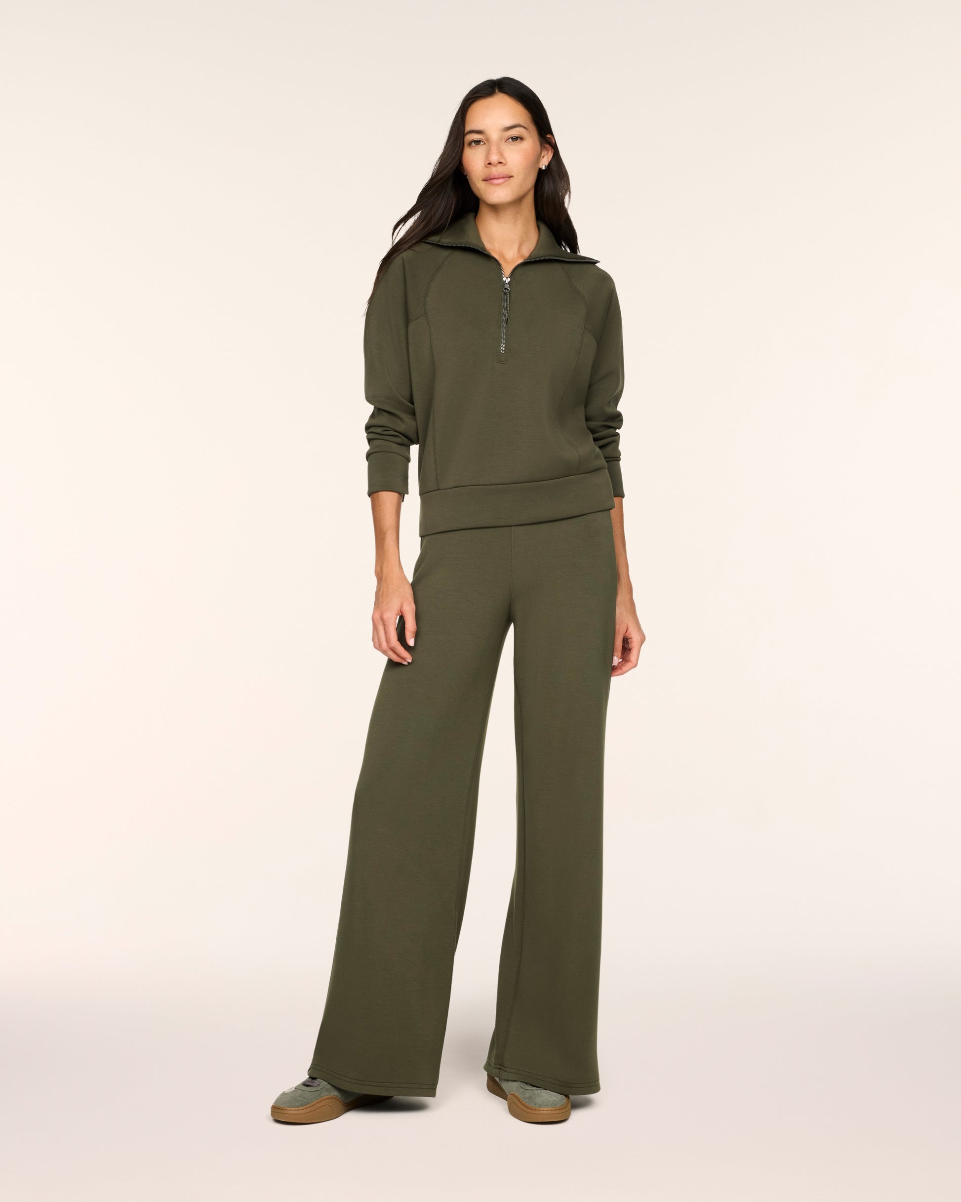 A woman modeling a matching olive green lounge set consisting of a half-zip long-sleeve top and wide-leg pants, photographed against a solid off-white studio background.