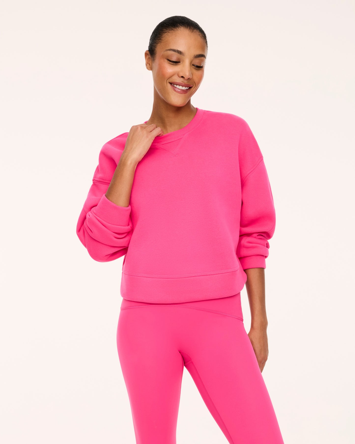 A woman modeling a bright pink crewneck sweatshirt and matching pink leggings against a clean off-white studio background.