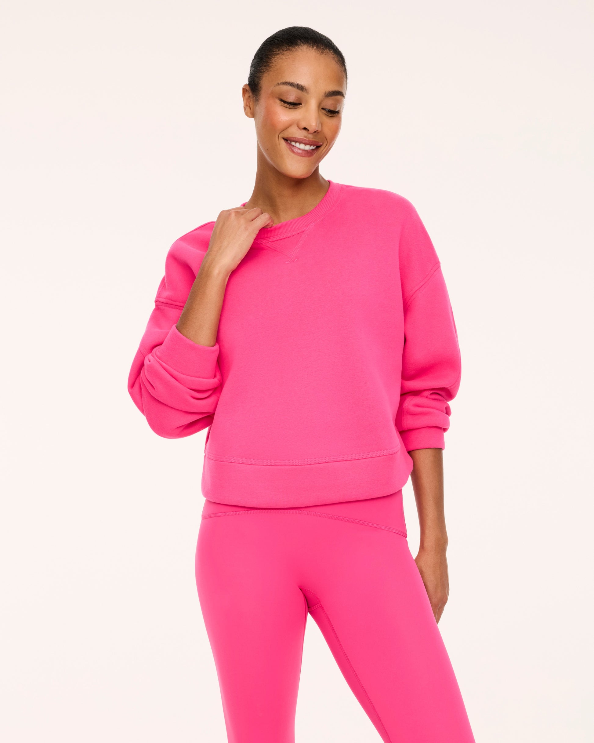 A woman modeling a bright pink crewneck sweatshirt and matching pink leggings against a clean off-white studio background.