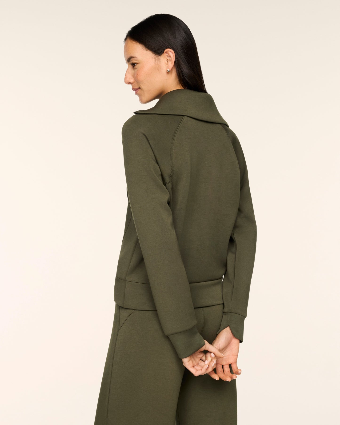 A woman shown from the back wearing an olive green long-sleeve coat with a wide collar and matching trousers, photographed against a plain light beige studio background.