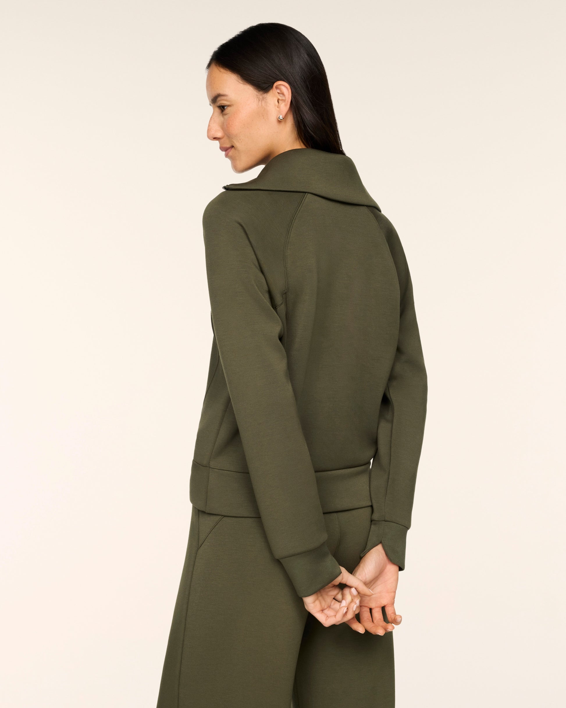A woman shown from the back wearing an olive green long-sleeve coat with a wide collar and matching trousers, photographed against a plain light beige studio background.