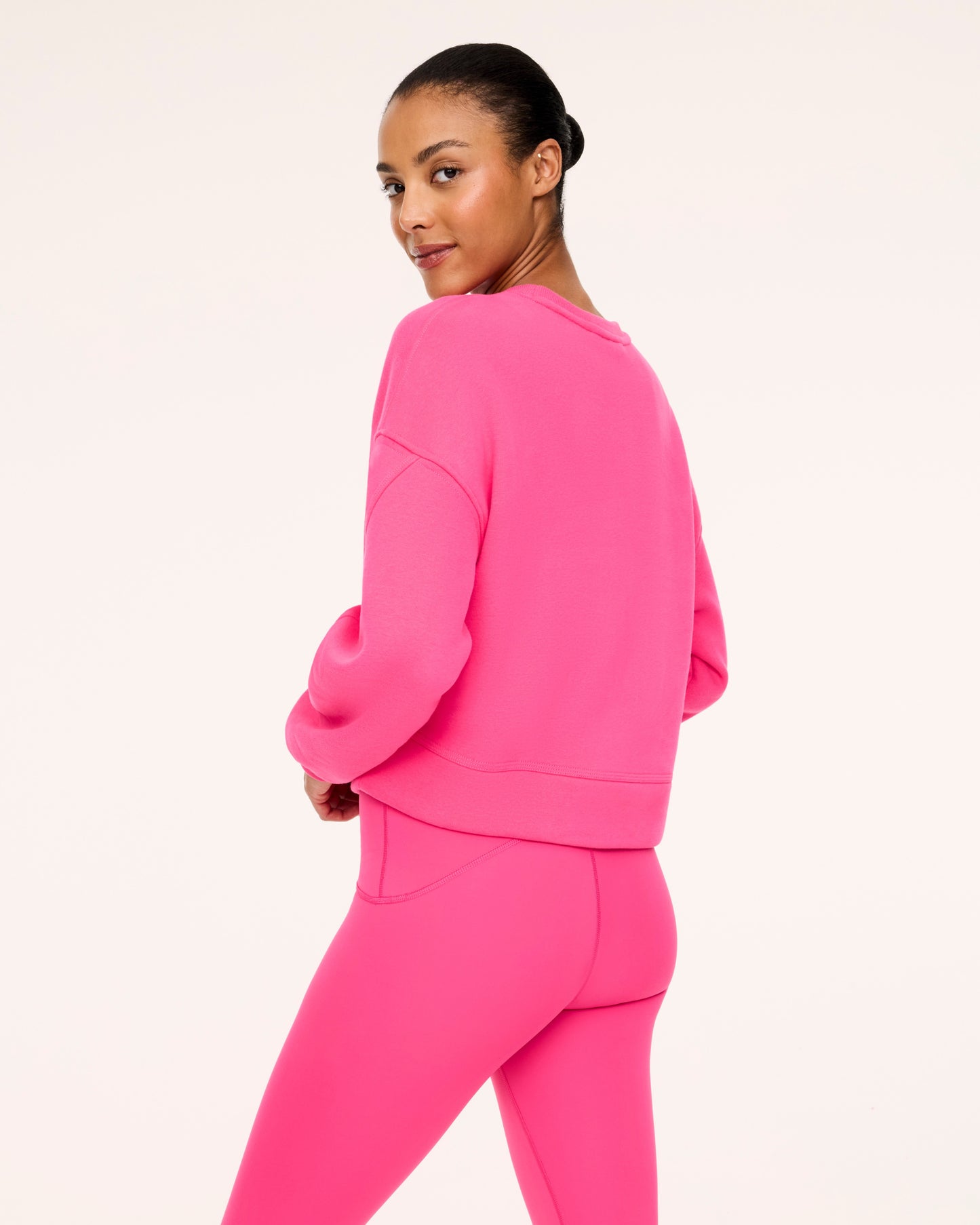 A woman modeling a bright pink matching lounge set consisting of a cropped long-sleeve sweatshirt and high-waisted leggings, photographed against a clean off-white studio background.