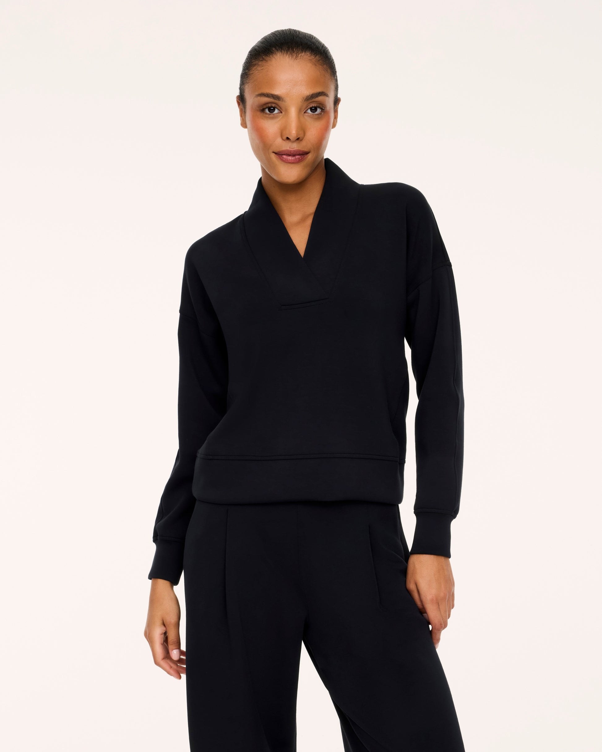A model wearing a black long-sleeve top with a unique folded collar and matching black trousers, photographed against a solid off-white studio background.