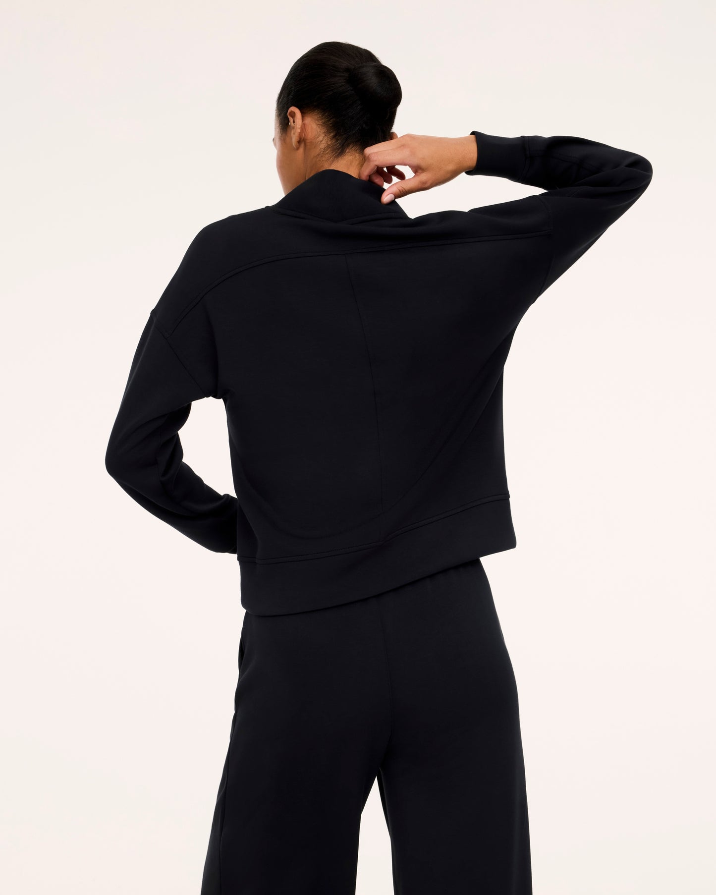 A model shown from the back wearing a black long-sleeve cropped zip-up top and matching black pants against a plain off-white studio background.